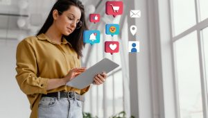 Simbolizando uma campanha promocional completa: ulher de óculos de grau usando tablet e, acima dele, estão ícones digitais coloridos como carrinho de compras, mão fazendo sinal de jóia (curtida), e-mail (contato), sino (notificações), emoji com corações nos olhos, ponto de localização, avatar de rede social e coração (curtida)
