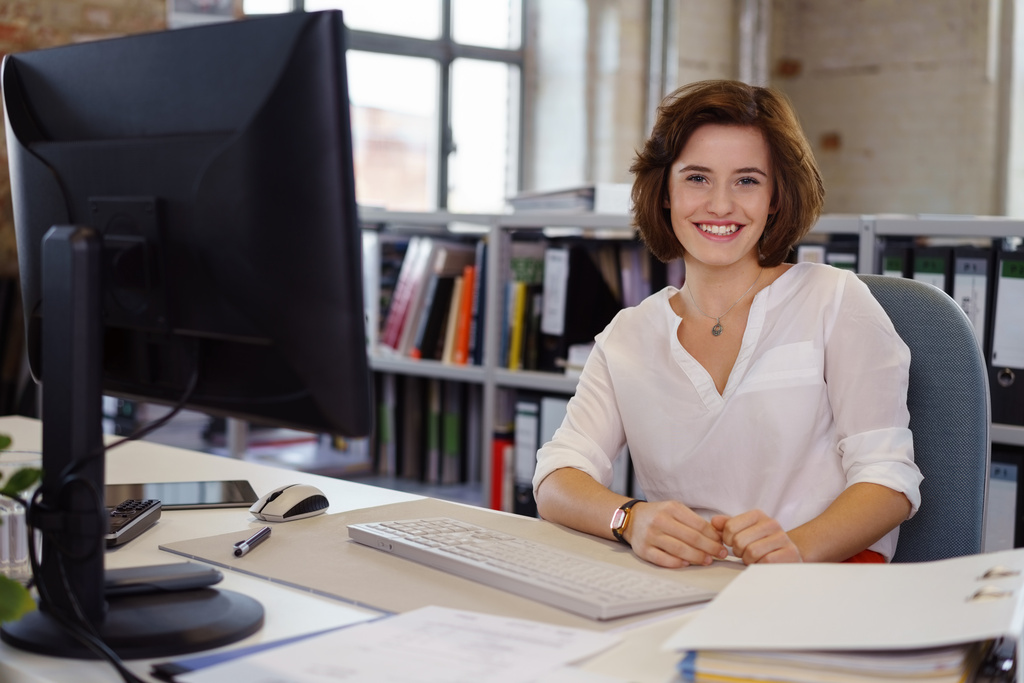 A mesma mulher da imagem anterior está sentada em sua mesa de trabalho, sorrindo diretamente para a câmera. À sua frente, há um monitor de computador, teclado e papéis organizados. O ambiente de escritório ao fundo é amplo, com janelas grandes e prateleiras repletas de pastas de arquivo.