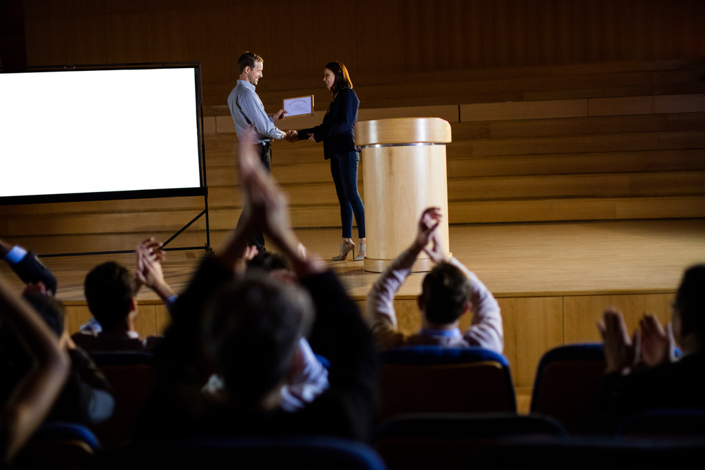 Executiva recendo um brinde tecnológico em um evento, plateia aplaudindo.