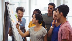 Mulher escrevendo em quadro branco, com notas adesivas coloridas para equipe corporativa, e equipe atenta ao quadro.