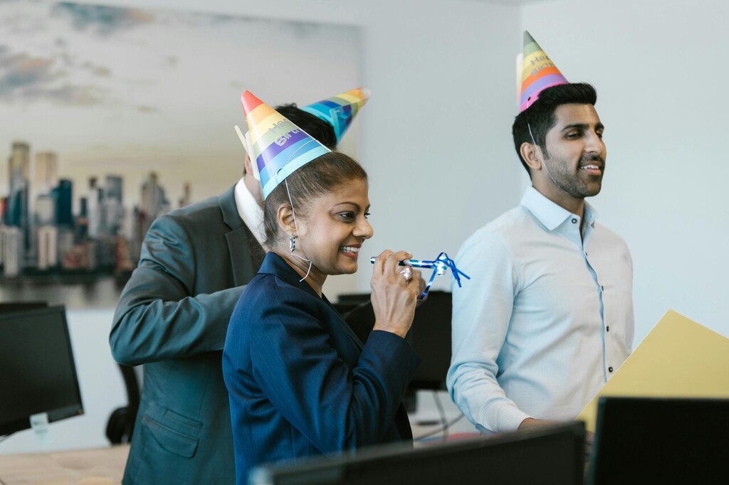Pessoas no escritório usando chapéus de festa e assoprando adereços em celebração de aniversário de empresa.