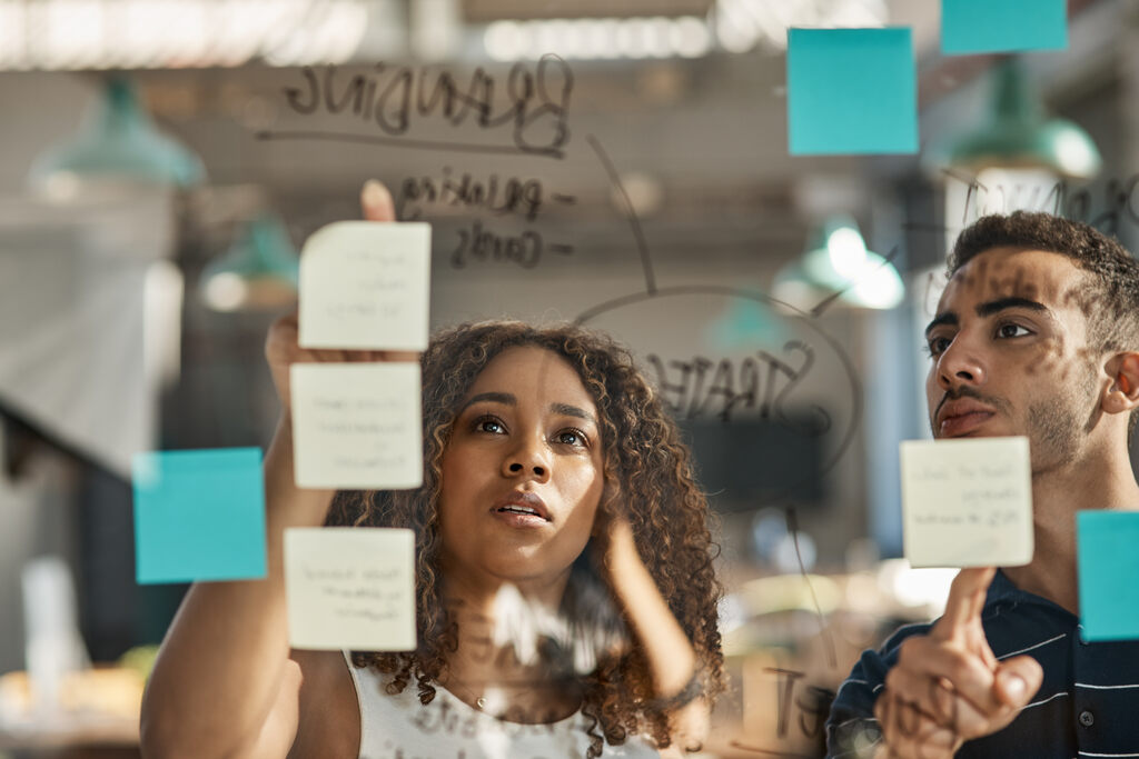 Dois profissionais, uma mulher negra com cabelos cacheados e um homem jovem, olham atentamente para uma parede de vidro transparente em um escritório moderno. Eles estão organizando post-its coloridos e escreveram a palavra "Branding" no vidro com um marcador. A cena foca na expressão de concentração e planejamento estratégico, ilustrando um processo de brainstorming e estruturação de marca.