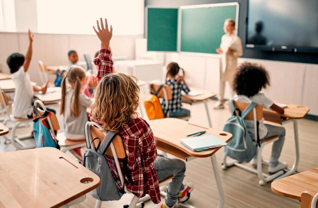 Vista do fundo de uma sala de aula iluminada onde vários alunos estão sentados em suas carteiras. Um aluno em destaque, vestindo uma camisa xadrez vermelha, está com a mão levantada para participar da aula. Ao fundo, a professora está em pé próxima ao quadro verde e a uma tela digital.