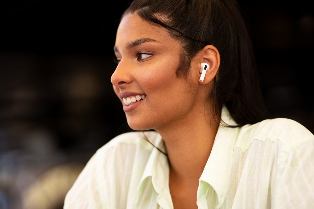 Mulher jovem usando fones de ouvido sem fio, sorrindo e olhando para o lado, em ambiente interior bem iluminado.